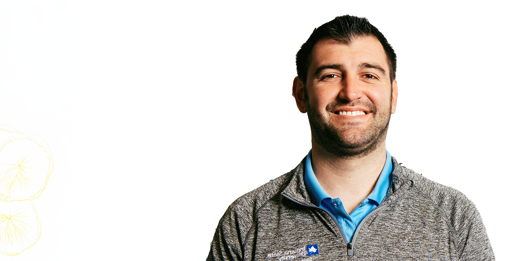 A white male with short black hair with a matching mustache and beard poses before a white backdrop. He wears a grey Indiana State University zipped-up jacket with a blue undershirt.